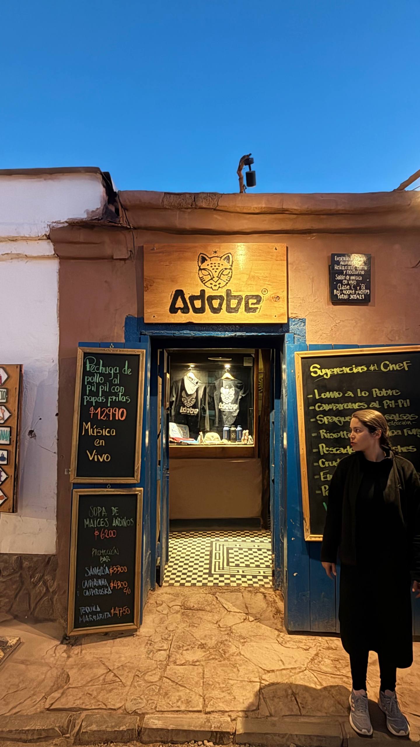 Fachada do restaurante Adobe em San Pedro de Atacama: paredes de adobe cor de terra, molduras azuis, o logo do puma e lousas de menu na porta