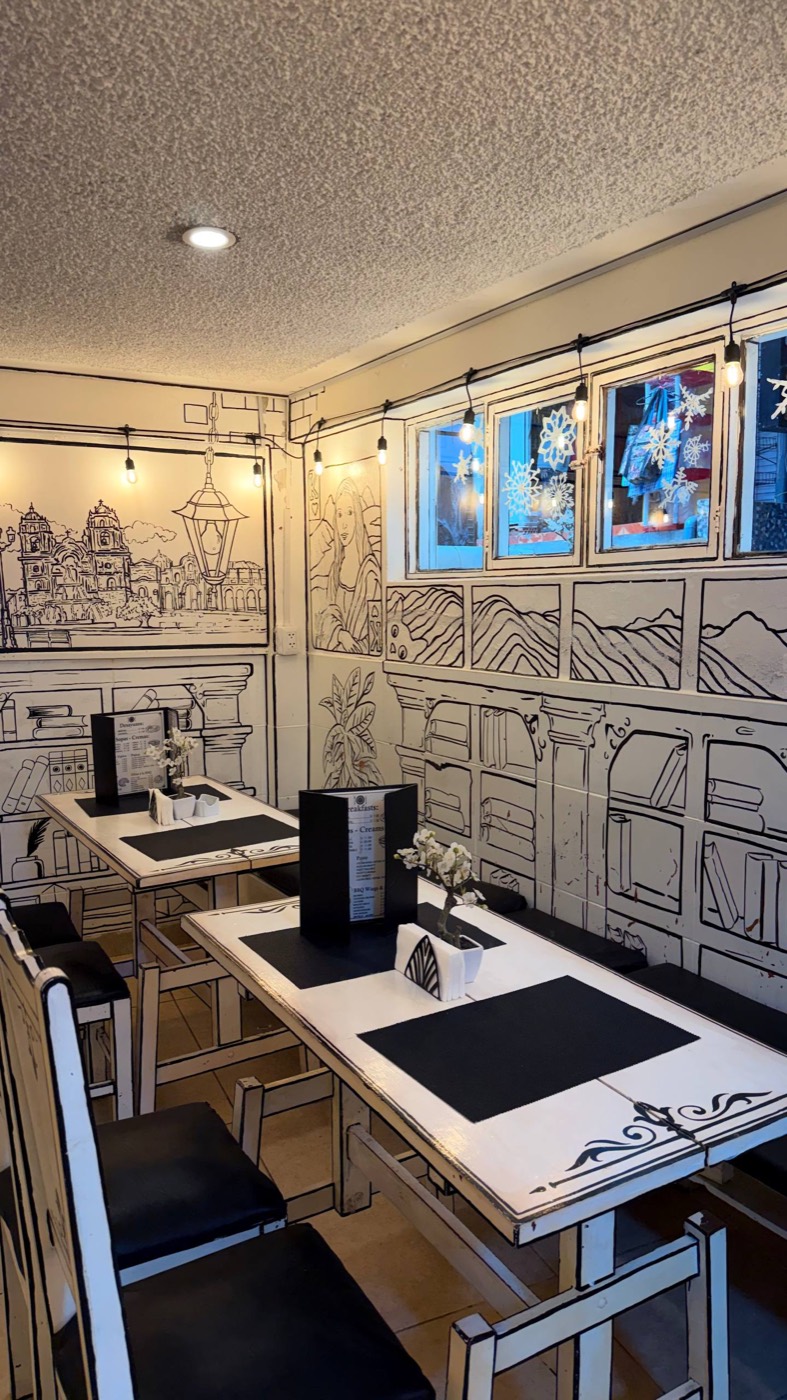 O salão do Art Café Machupicchu: paredes, mesas e cadeiras brancas com tudo desenhado em traço preto, como um caderno de colorir em tamanho real, com estantes, uma catedral e uma Mona Lisa rabiscadas na parede e luzinhas amarelas no teto