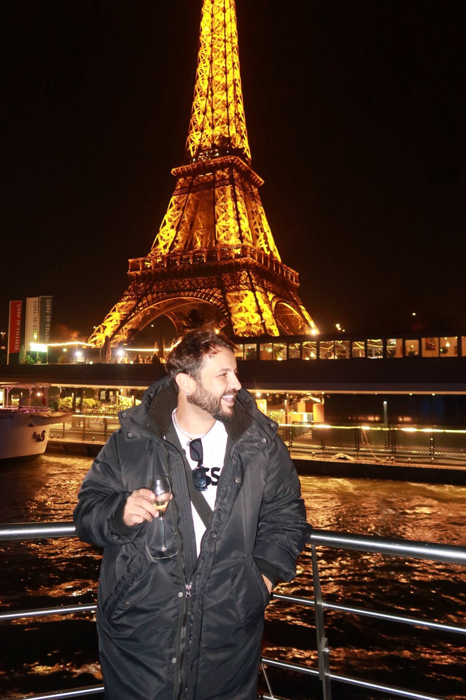 O Dan de taça na mão no barco pelo Sena, com a Torre Eiffel iluminada atrás