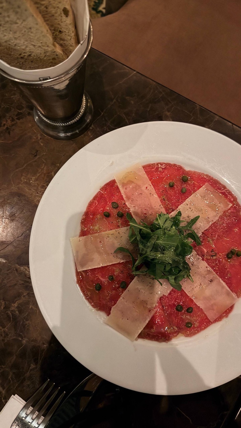 Carpaccio de res do Bottega Dasso: lâminas finas de carne com lascas de parmesão em cruz, alcaparras e rúcula no centro, num prato branco sobre mesa de mármore escuro, com a cesta de pão ao lado