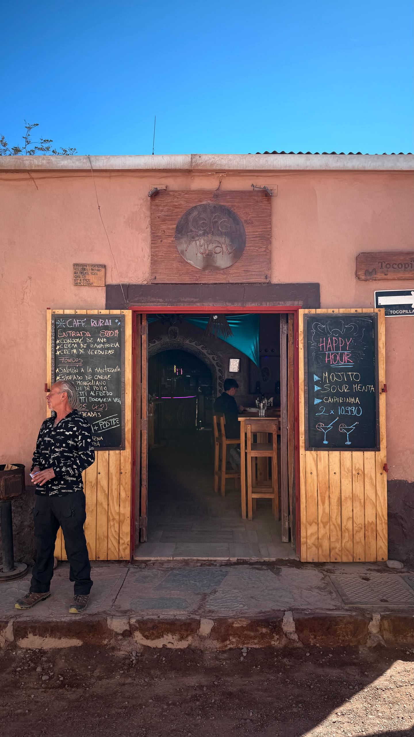 Fachada de adobe do Café Rural em San Pedro de Atacama, com lousas de menu e happy hour na porta e o interior escuro de bar ao fundo