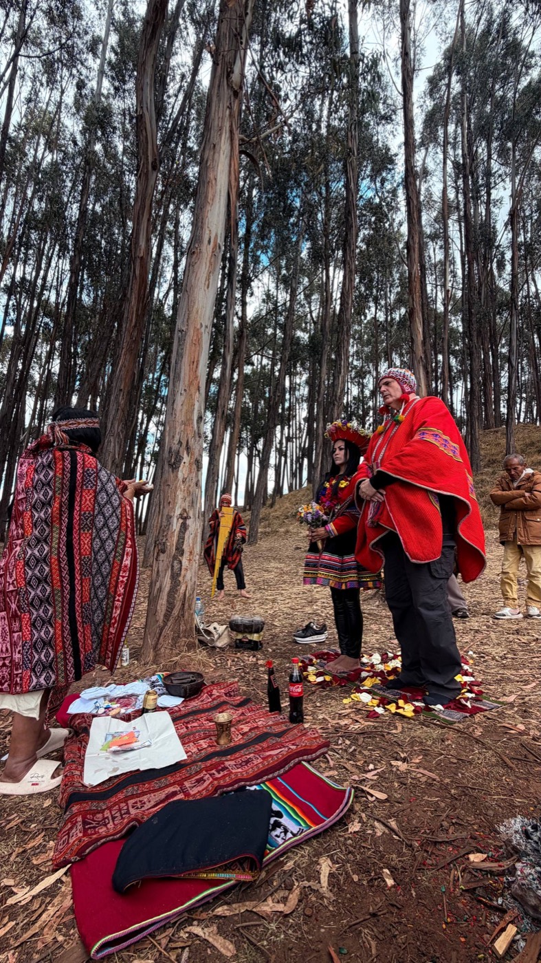Casamento andino num bosque de eucaliptos perto de Cusco: a oficiante de poncho vermelho e preto de costas, a noiva com coroa de flores e o noivo de poncho vermelho e chullo em pé sobre pétalas, um músico de flauta ao fundo e a mesa da oferenda estendida no chão