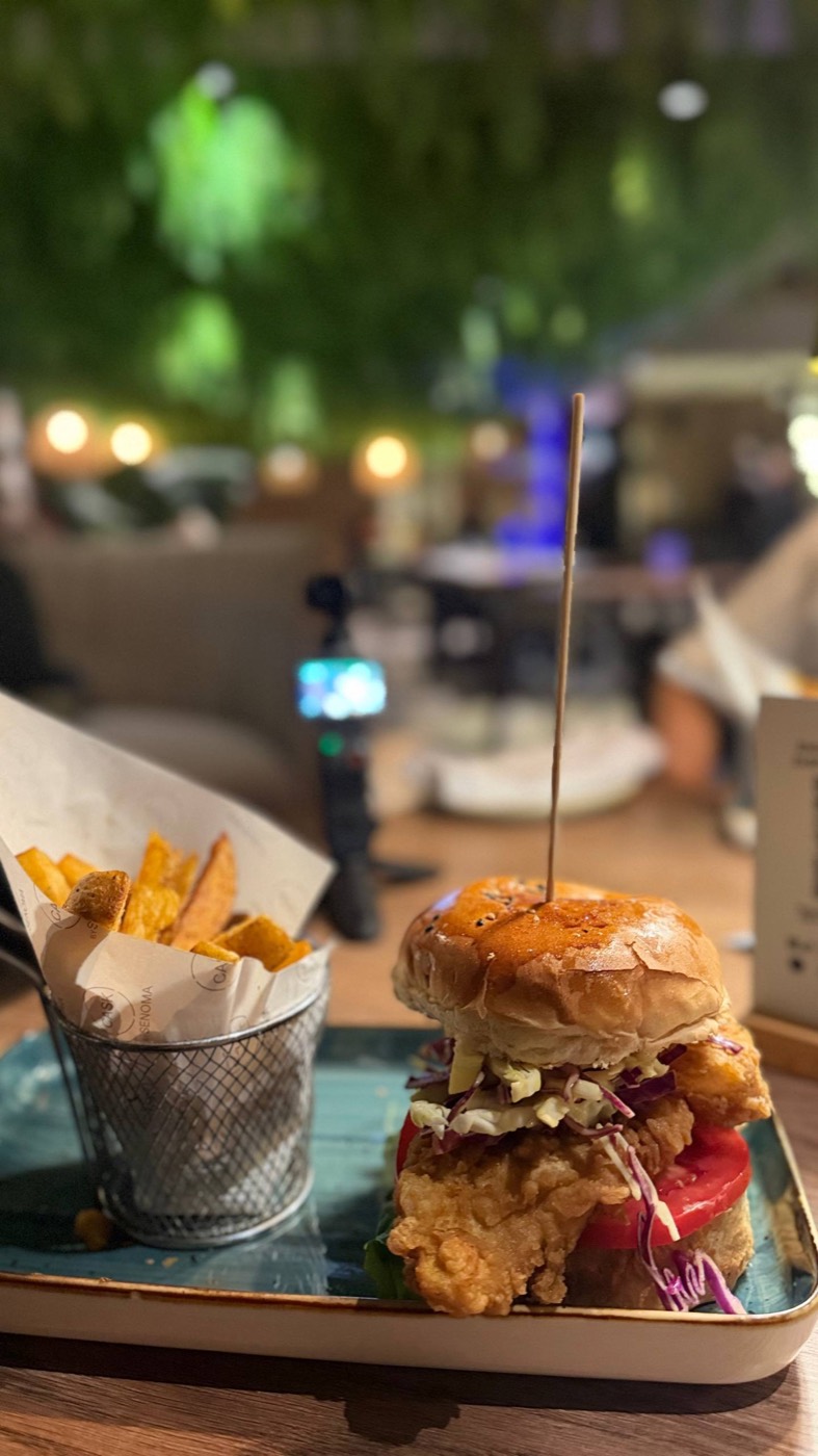 Crispy Chicken Sandwich do Cask: frango empanado alto no pão brioche com coleslaw roxo e tomate, espetado com um palito, e a cestinha de metal com batatas rústicas ao lado, com o bar iluminado ao fundo