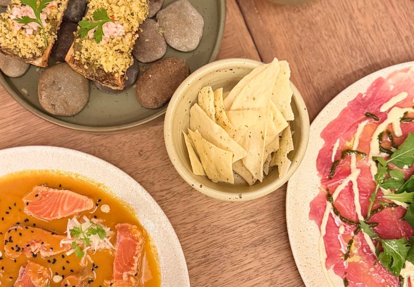 Mesa do Chicha em Cusco vista de cima: tuétano gratinado servido sobre pedras, tiradito de truta em molho alaranjado com gergelim preto e um carpaccio de alpaca com rúcula, mais uma cumbuca de pão fino crocante