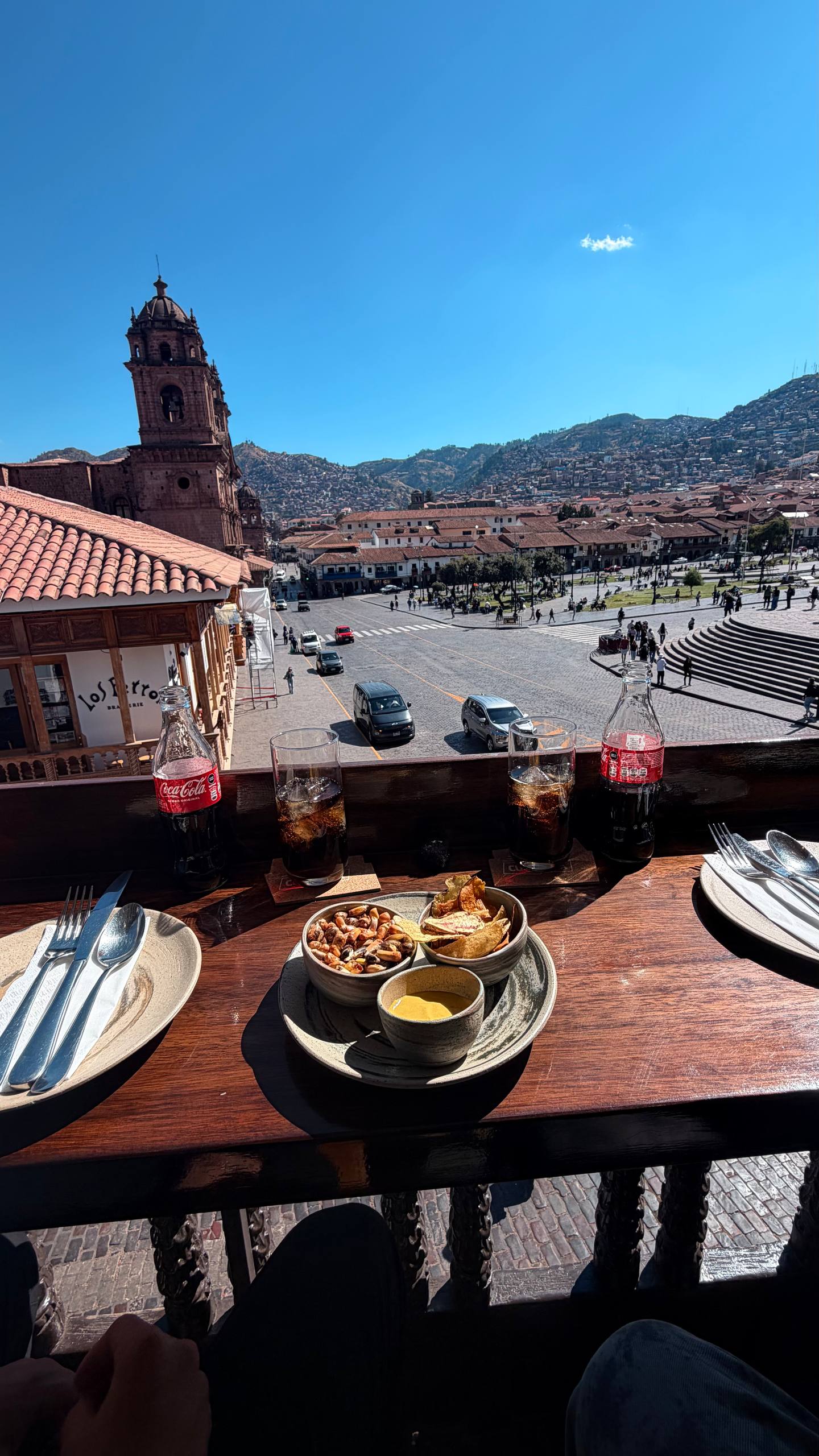 A vista da terraça do Casa Cusqueña: a Plaza de Armas de Cusco lá embaixo com a igreja, os telhados vermelhos e as montanhas ao fundo, e na mesa a entradinha de cortesia (chips, canchita e um molho) com as Coca-Colas