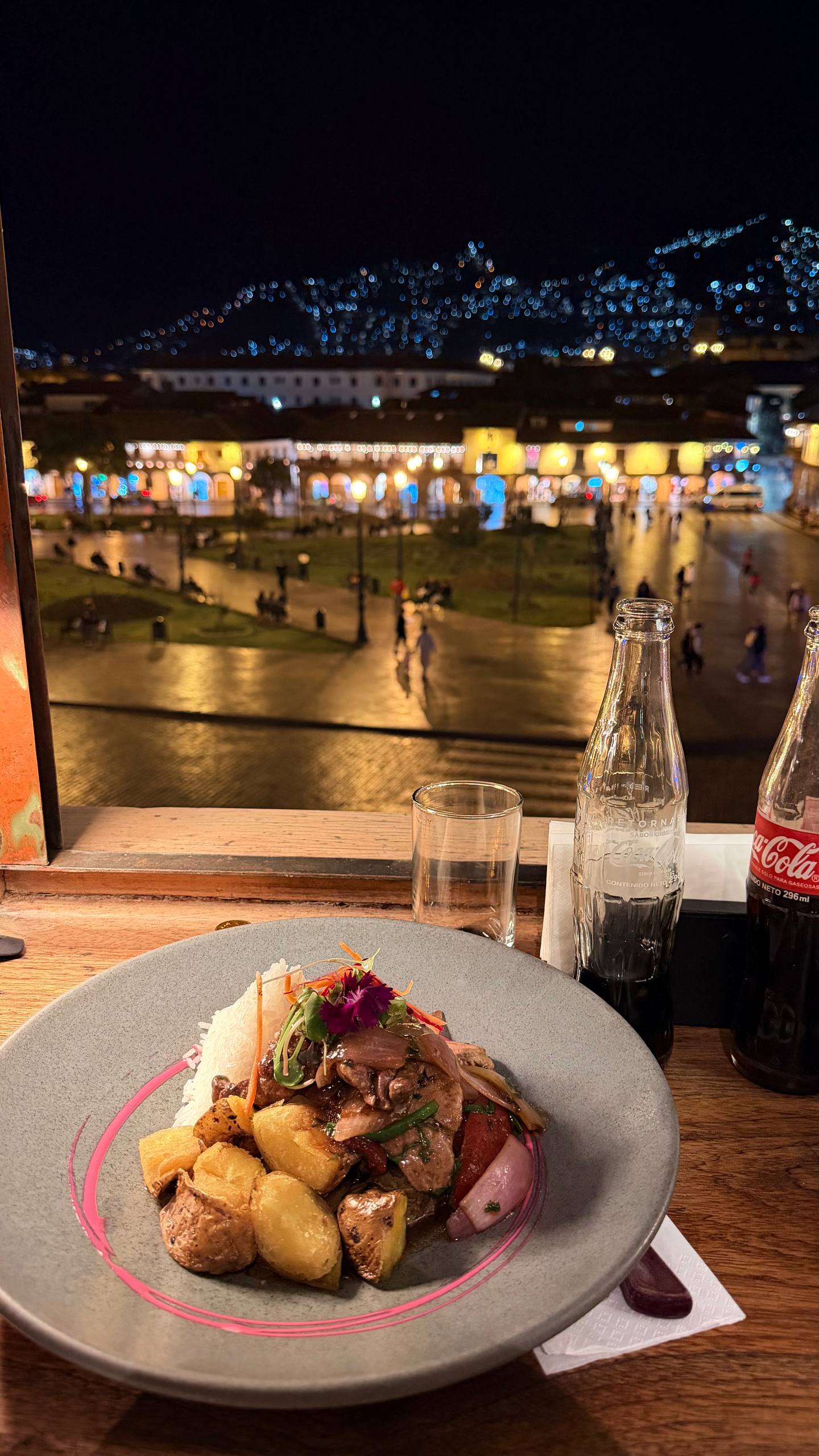 Meu prato de alpaca saltada no Chise: tiras de alpaca refogadas com cebola roxa, tomate e batata dourada, arroz branco e uma flor comestível por cima, com a Plaza de Armas de Cusco iluminada à noite ao fundo pela janela e as garrafinhas de Coca-Cola de vidro na mesa