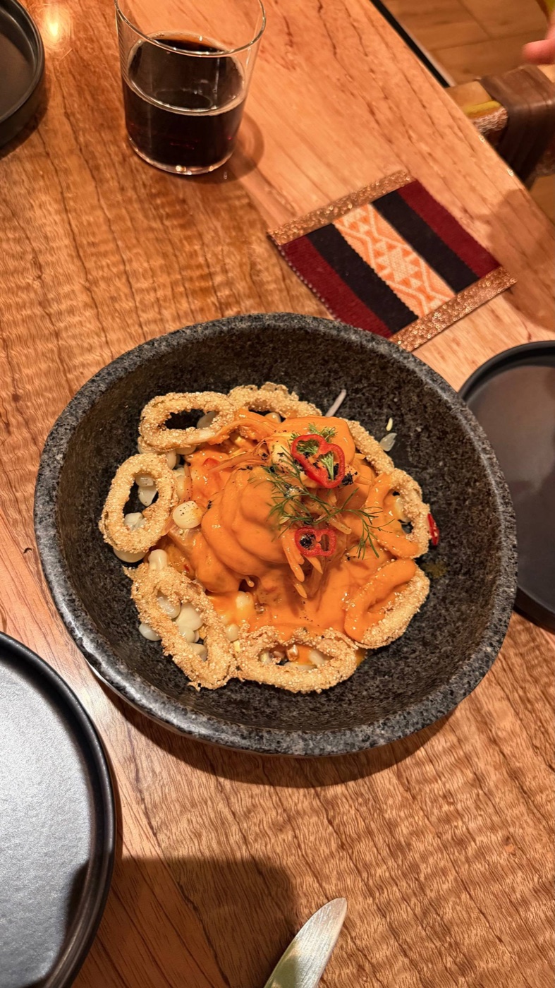 O ceviche do Morena numa tigela de pedra: leche de tigre alaranjado, aros de lula empanados em volta, choclo e pimenta por cima, na mesa de madeira