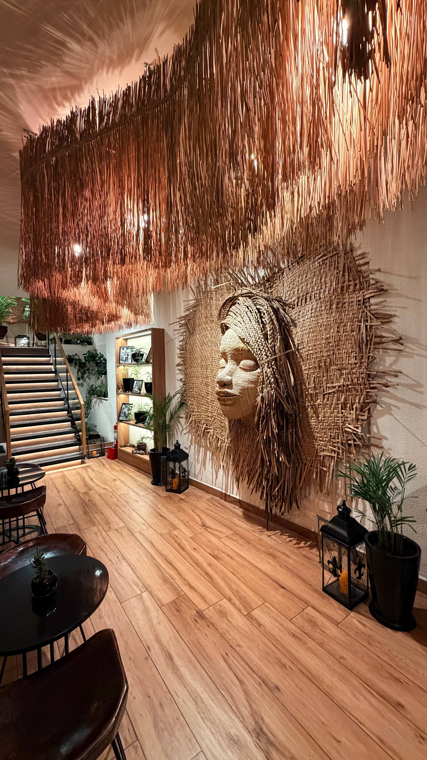 O salão de entrada do Morena: uma escultura enorme de um rosto de mulher andina feito em fibra natural trançada na parede, teto de palha pendente, iluminação quente, plantas e uma escada de madeira ao fundo
