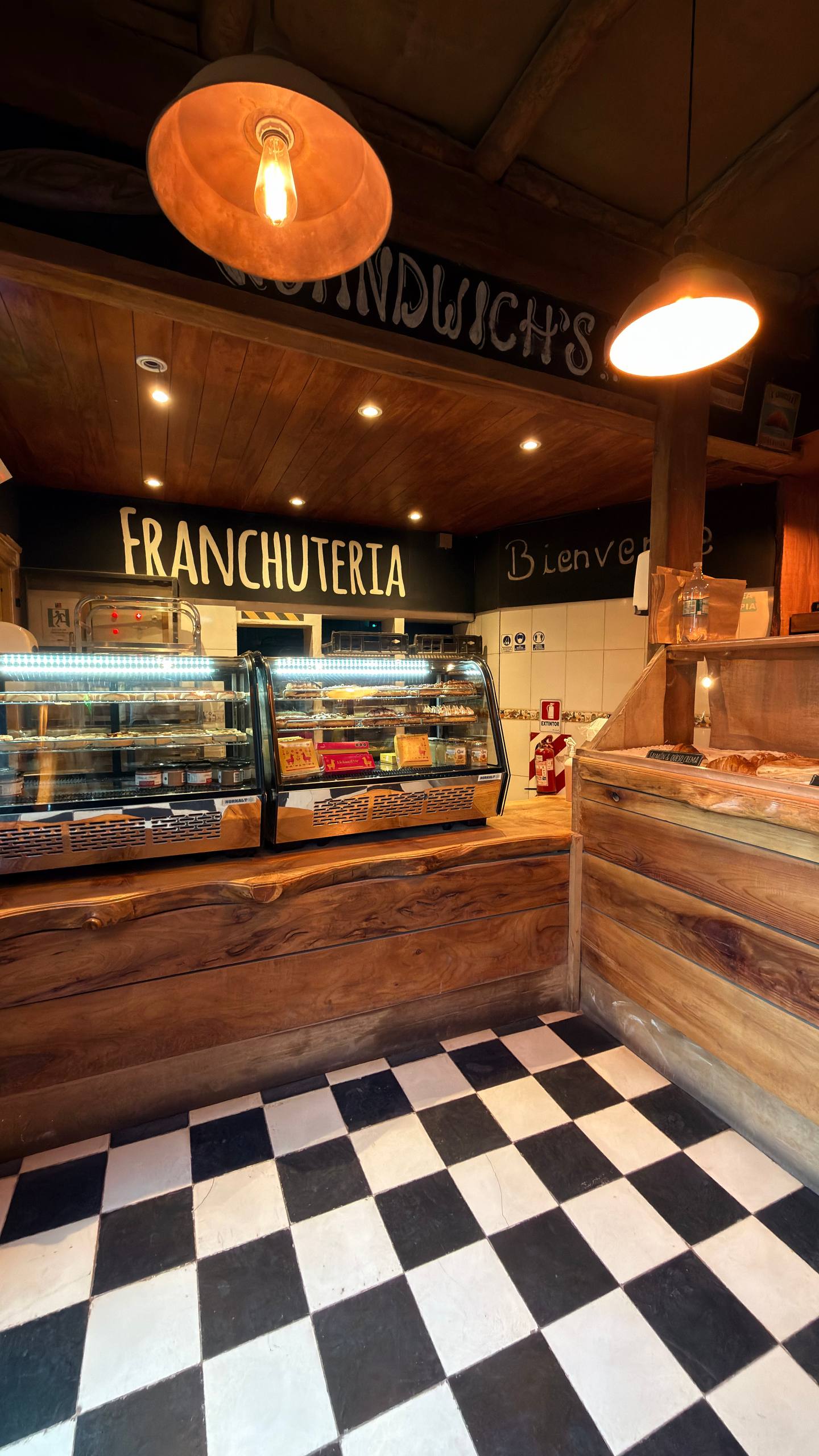 Interior da Franchuteria: 'Bienvenue' pintado na parede, vitrines de doces, chão xadrez preto e branco e luz quente