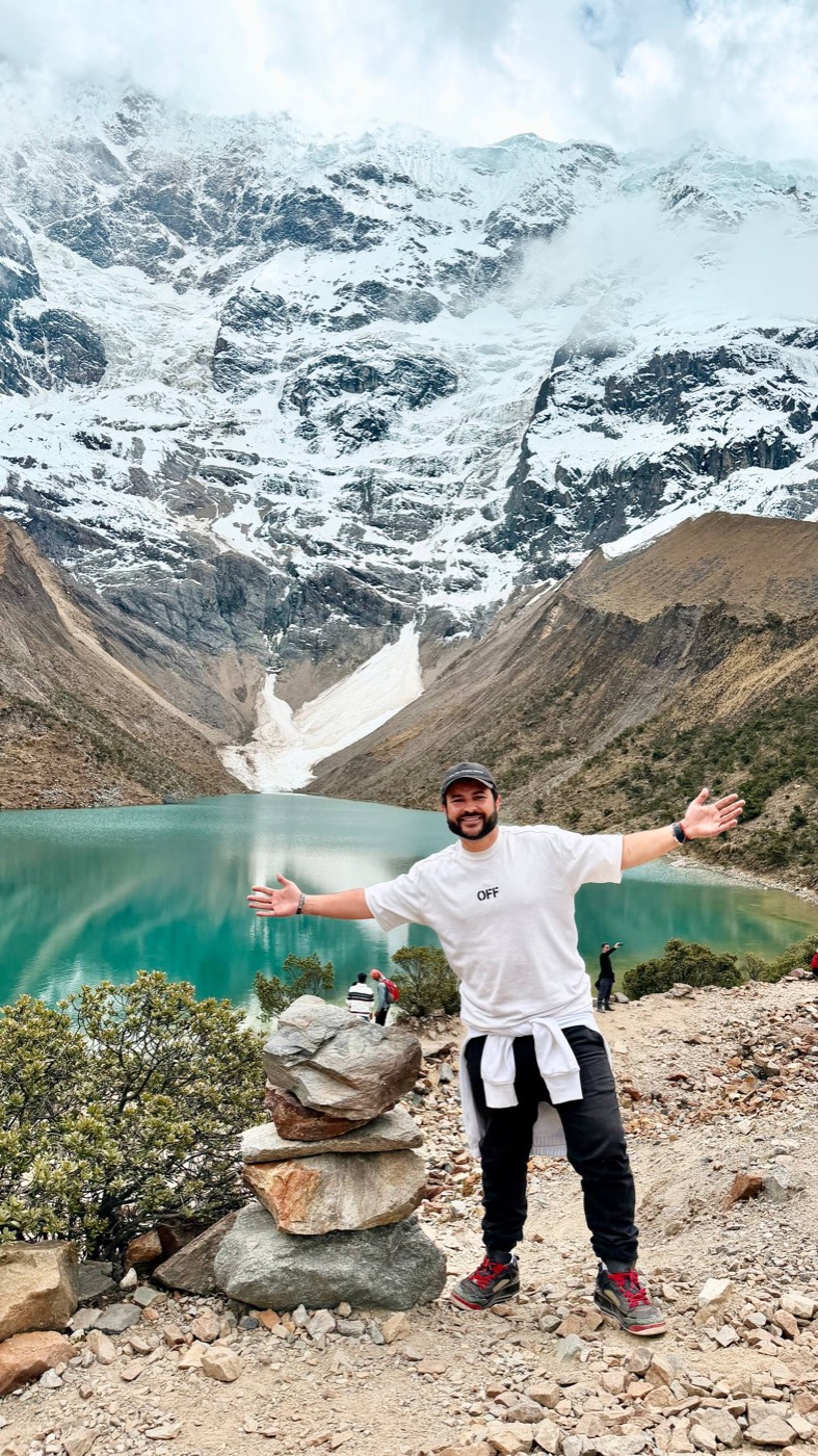 Dan de braços abertos, boné e camiseta branca, na beira da Laguna Humantay: a lagoa de água turquesa de degelo, o paredão do glaciar do Salkantay todo nevado atrás, e uma pilha de pedras empilhadas ao lado dele