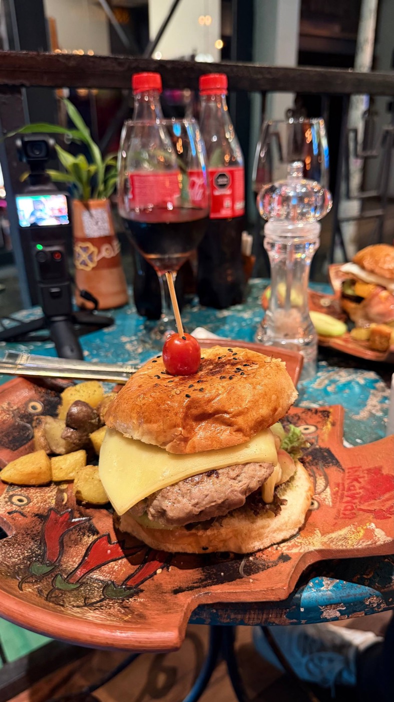 O hambúrguer de alpaca do Inka Wasi: pão brioche com gergelim e uma cereja espetada no topo, fatia de queijo andino derretendo sobre o hambúrguer de alpaca grelhado, servido num prato de cerâmica em formato de chakana com o nome Inkawasi pintado, com batatas nativas douradas ao lado
