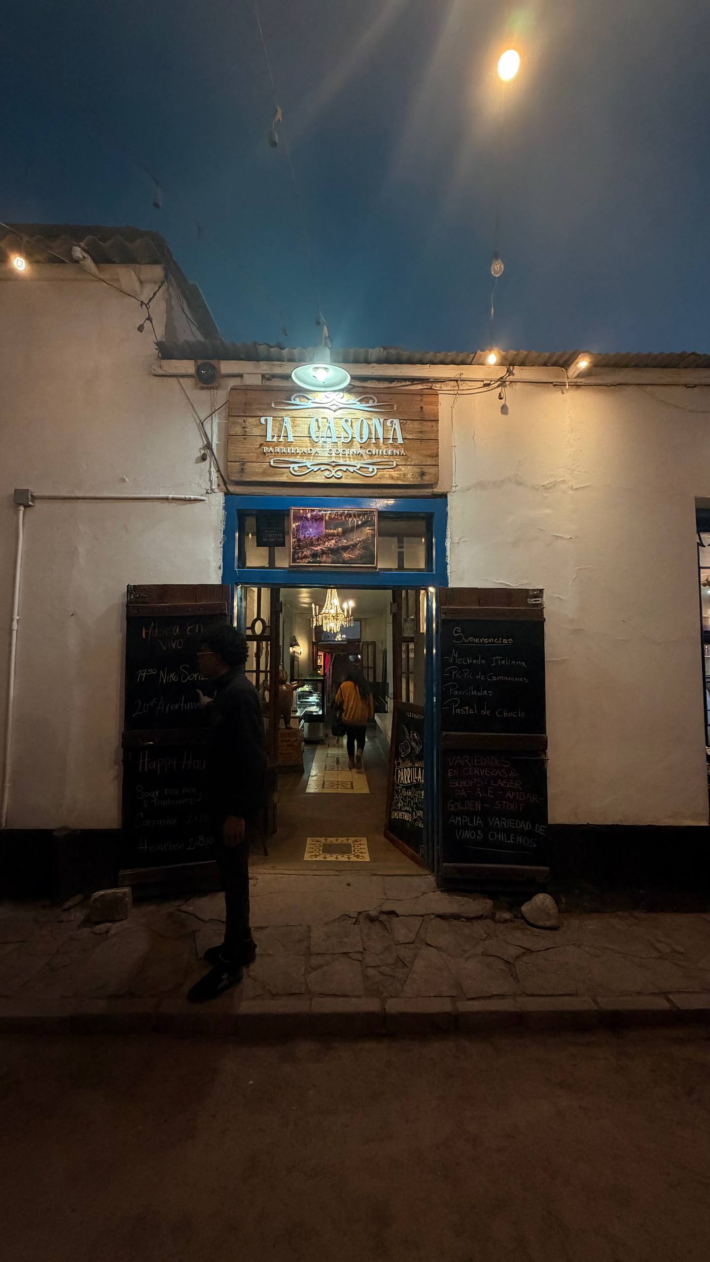 Fachada noturna do La Casona em San Pedro de Atacama, com o letreiro de madeira 'Parrillada y Cocina Chilena', lousas de menu e o lustre visível lá dentro