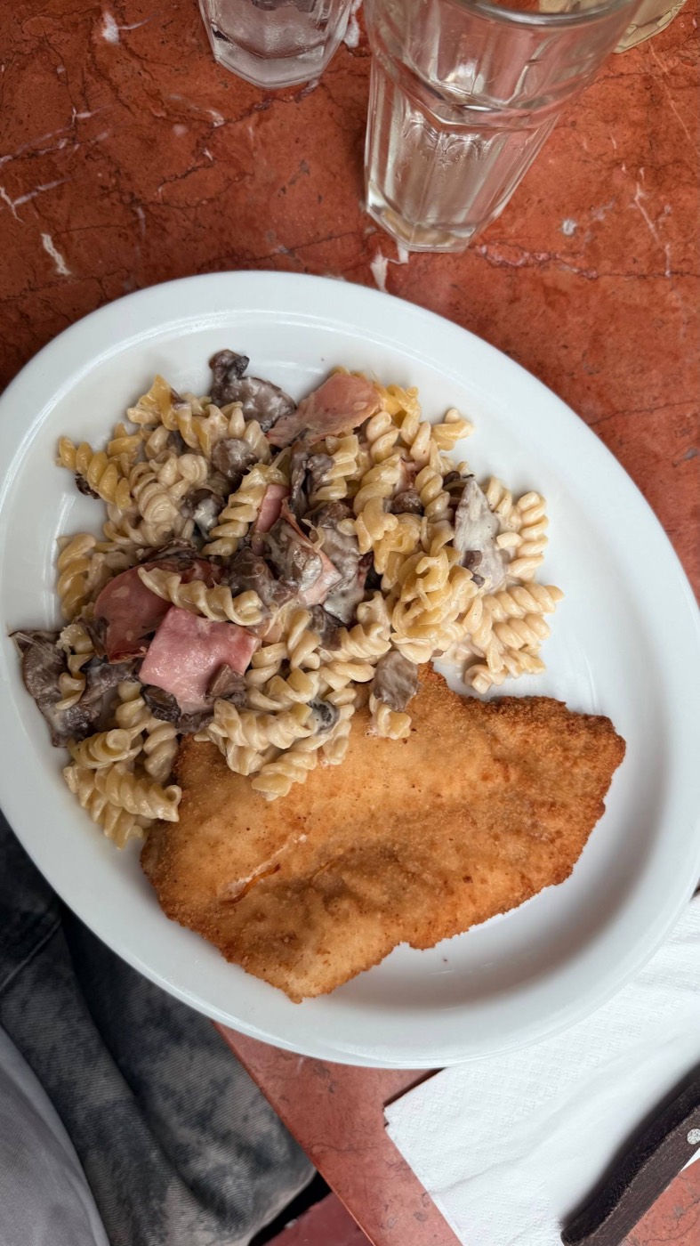 Milanesa de frango dourada ao lado de fusilli no molho Alfredo com champignons e presunto, num prato branco sobre mesa de mármore vermelho da La Bodega de la Trattoria