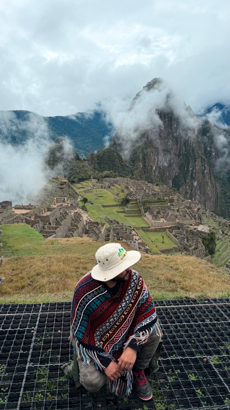 Dan de poncho na frente do Machu Picchu, com a cidadela de pedra e a montanha Huayna Picchu ao fundo, o céu abrindo entre as nuvens