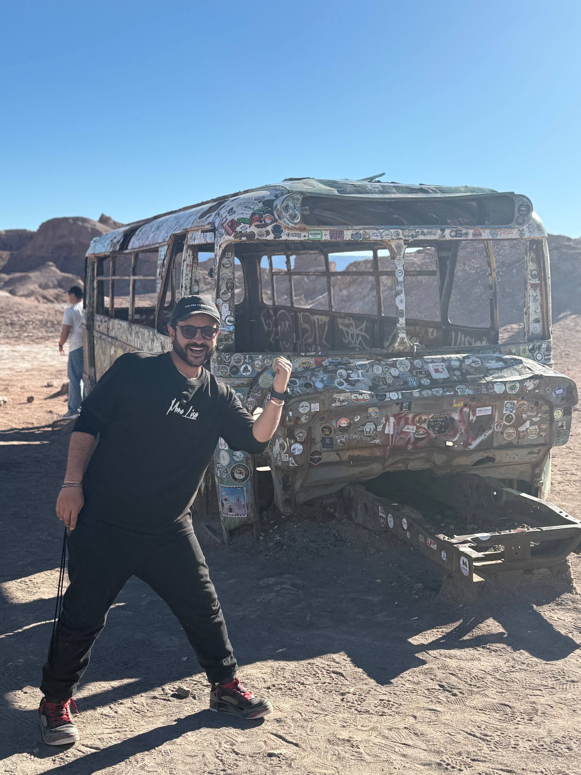 Dan posando de punho erguido ao lado do Magic Bus, o ônibus abandonado coberto de adesivos no meio do deserto do Atacama