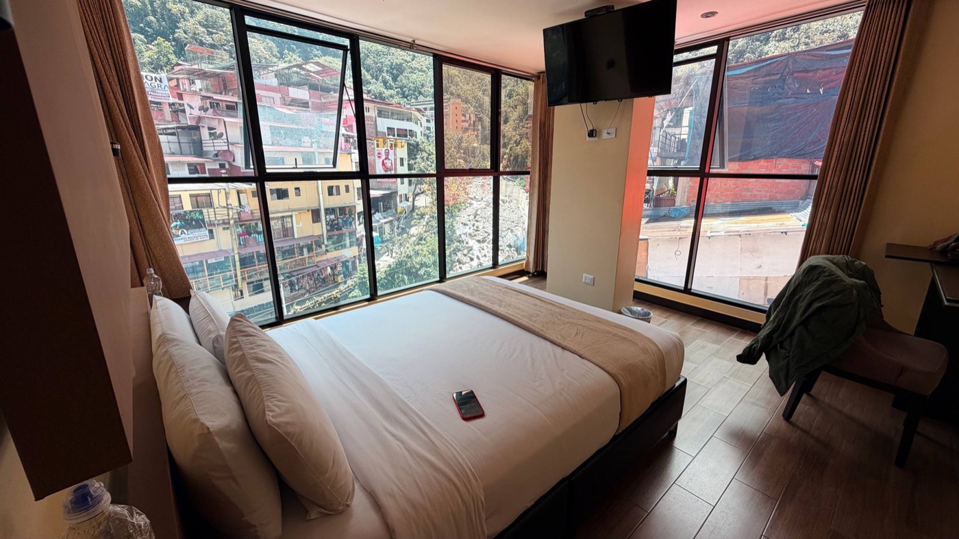 Quarto do Hotel Raíces em Águas Calientes: cama de casal com travesseiros brancos e manta bege, janelas de canto do chão ao teto com vista pras casas coloridas da cidade, a montanha verde e o rio de pedras lá embaixo, TV na parede e piso de madeira