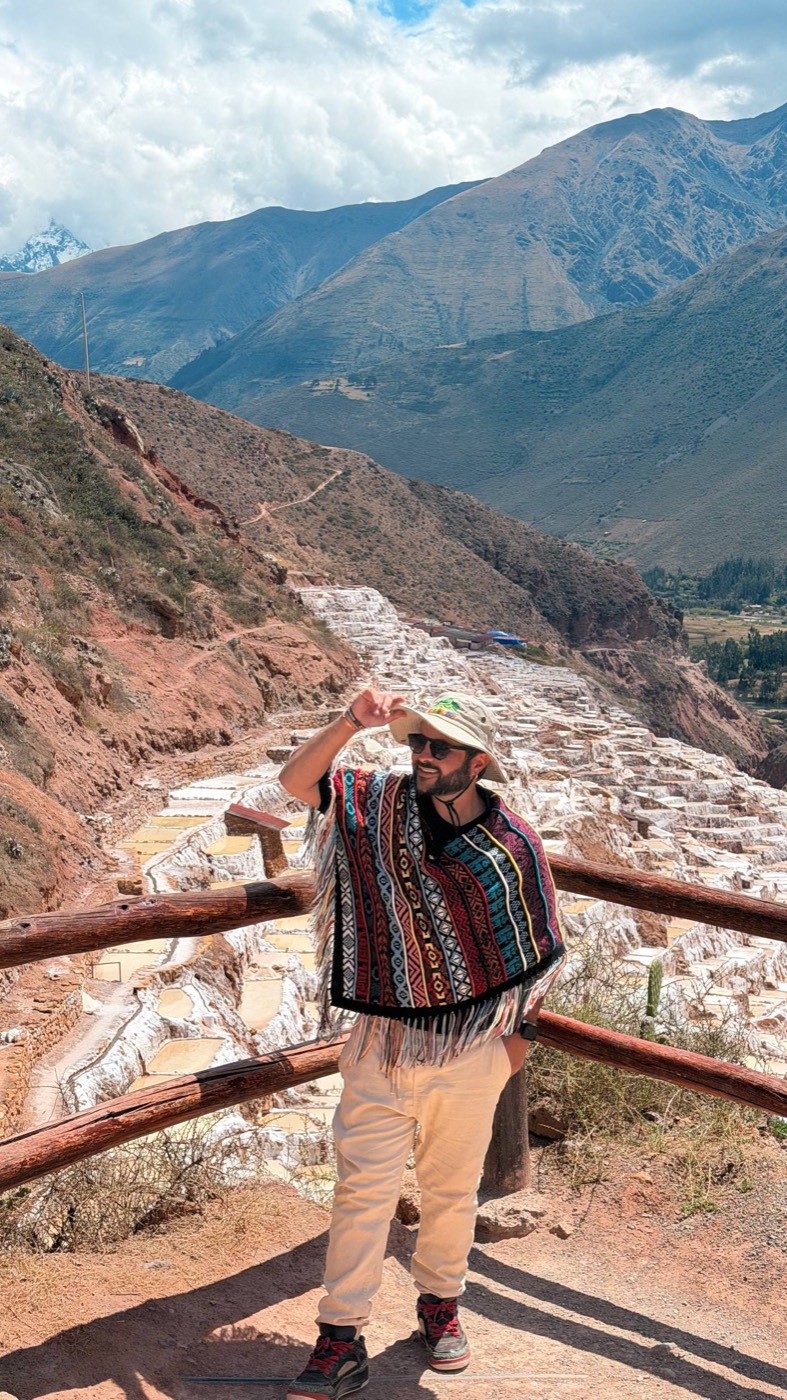 Dan de poncho andino e chapéu bucket, segurando a aba do chapéu, num mirante de madeira com as Salineras de Maras atrás: centenas de piscinas retangulares de sal branco descendo a encosta avermelhada da montanha, com o vale e picos ao fundo