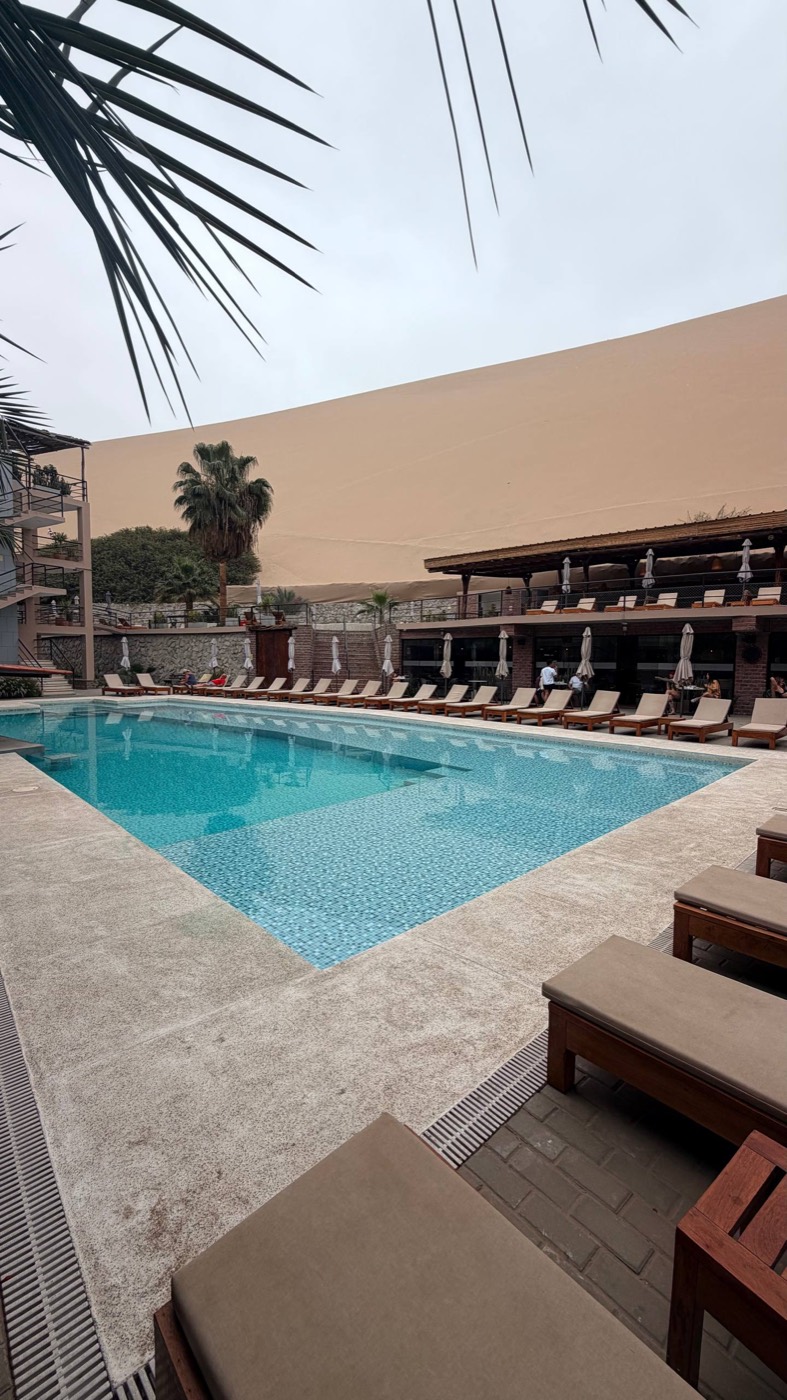 Piscina do Senoma Hotel em Huacachina, azul, com espreguiçadeiras de madeira em volta, palmeiras, e a duna gigante de areia subindo logo atrás do restaurante