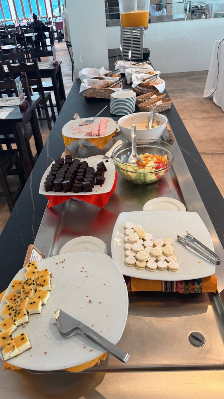 A mesa de sobremesas do buffet do Toto's House: alfajores com açúcar de confeiteiro, quadradinhos de cheesecake de maracujá, brownies, gelatina rosa, salada de frutas numa tigela de vidro e o suco na máquina ao fundo, com o salão de mesas de madeira atrás