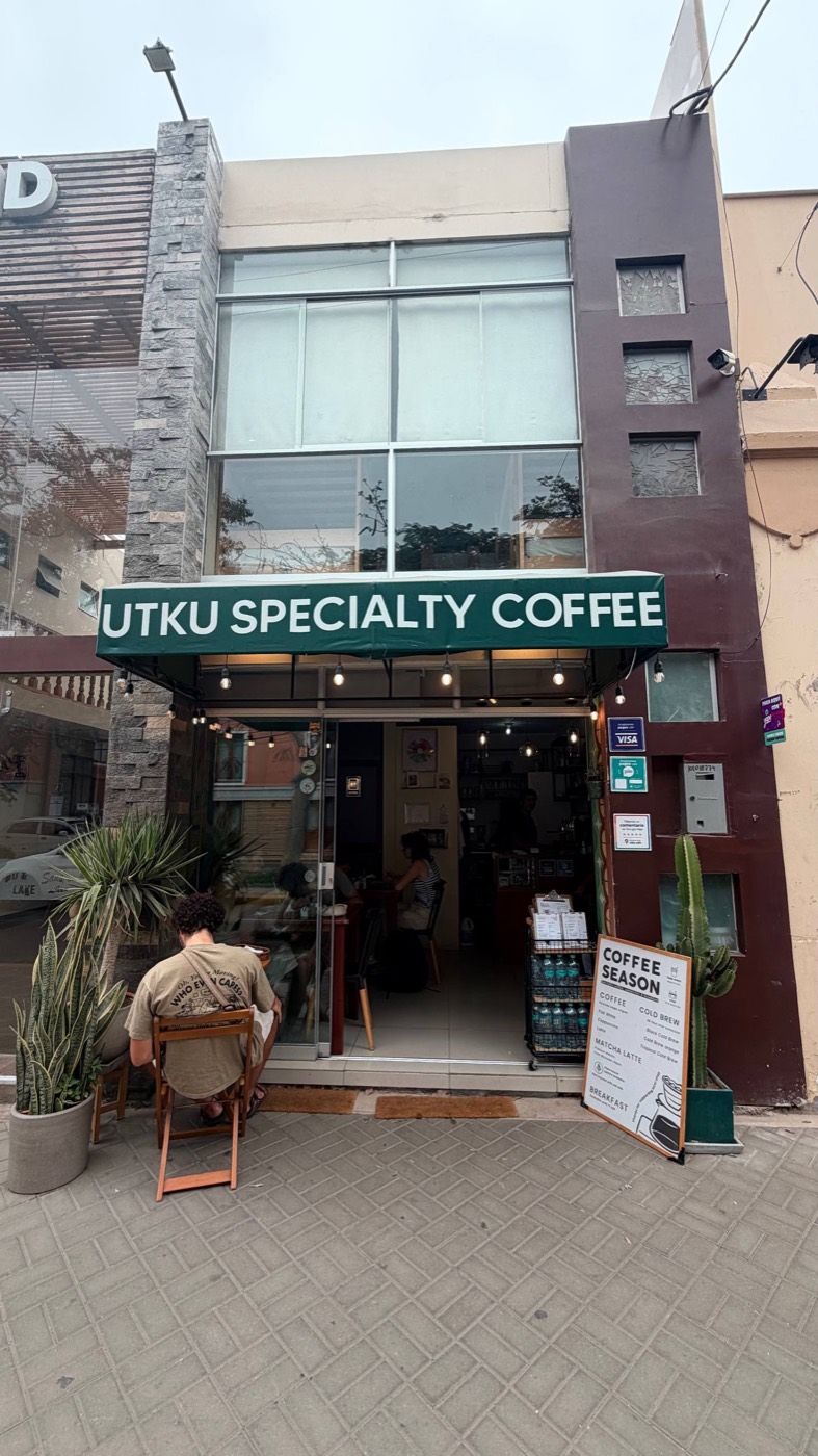 Fachada do Utku Specialty Coffee em Huacachina: toldo verde com o nome, porta de vidro aberta, luzinhas penduradas, cactos e plantas na calçada e uma pessoa sentada na mesinha de fora