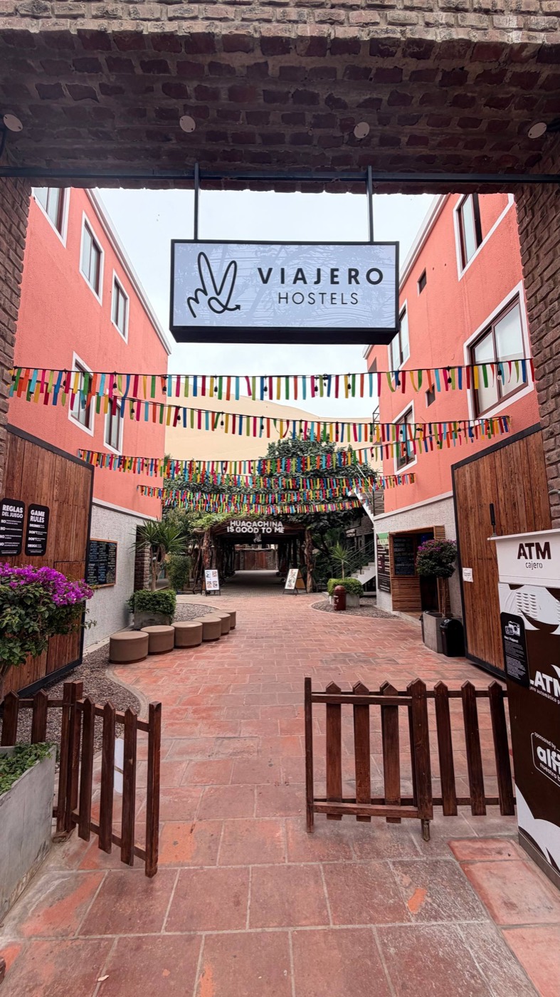 Entrada do Viajero Hostel em Huacachina: portão de tijolo, placa branca com o logo, bandeirinhas coloridas cruzando o pátio, a duna aparecendo no fundo e o letreiro Huacachina is good to me