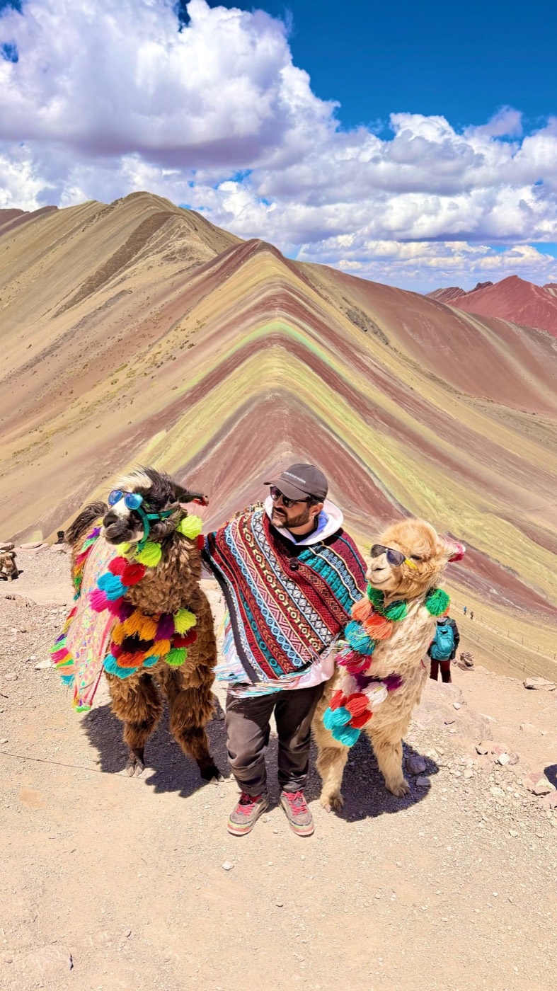 Dan de poncho andino colorido e boné, de óculos escuros, abraçado a duas lhamas enfeitadas com pompons coloridos e óculos escuros, no topo da Montanha de 7 Cores, com as faixas de terra colorida da montanha e o céu azul com nuvens atrás